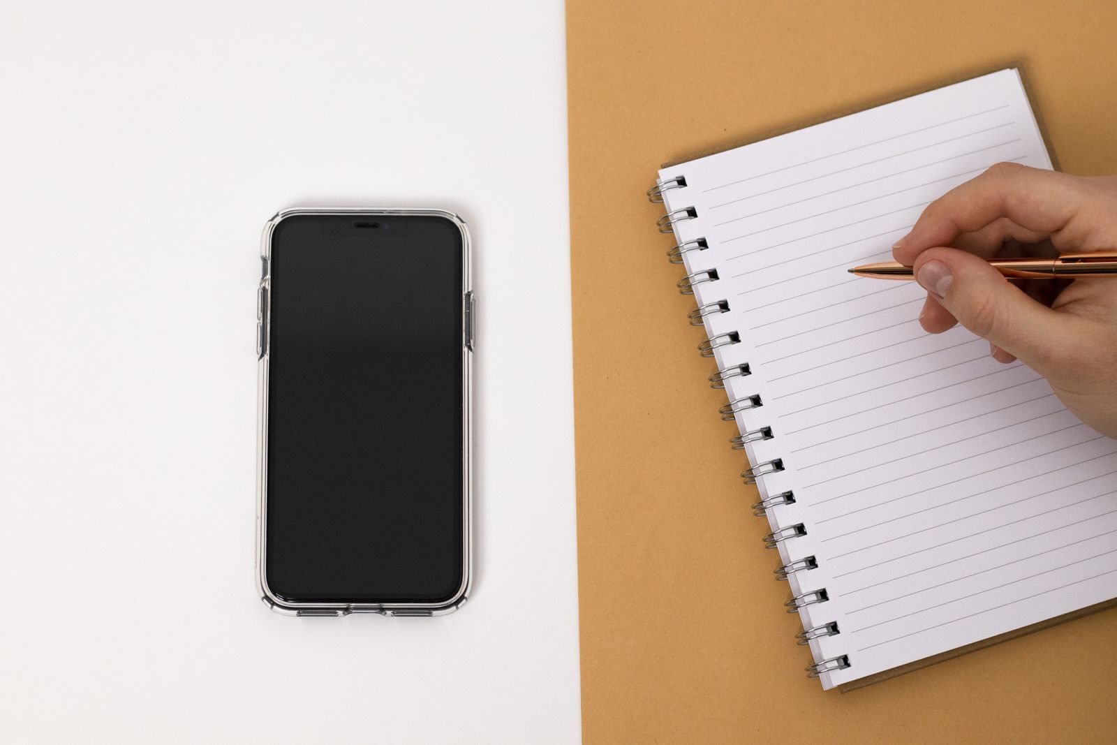 Pessoa faz anotações em um caderno ao lado de um celular.