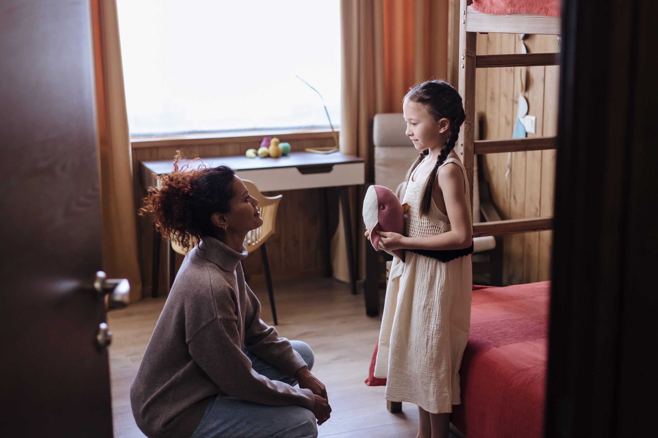Mulher agachada conversa com uma menina que segura uma almofada no quarto.