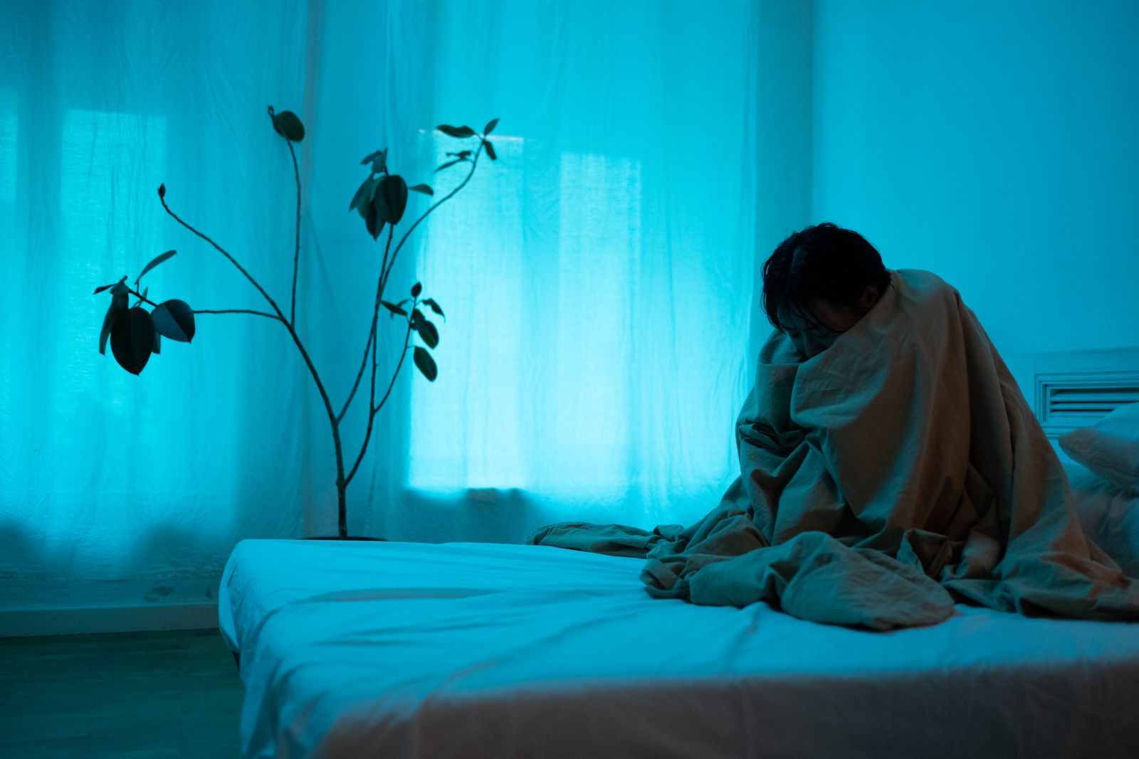 Mulher sentada na cama, envolta em um cobertor, em quarto com iluminação azulada.