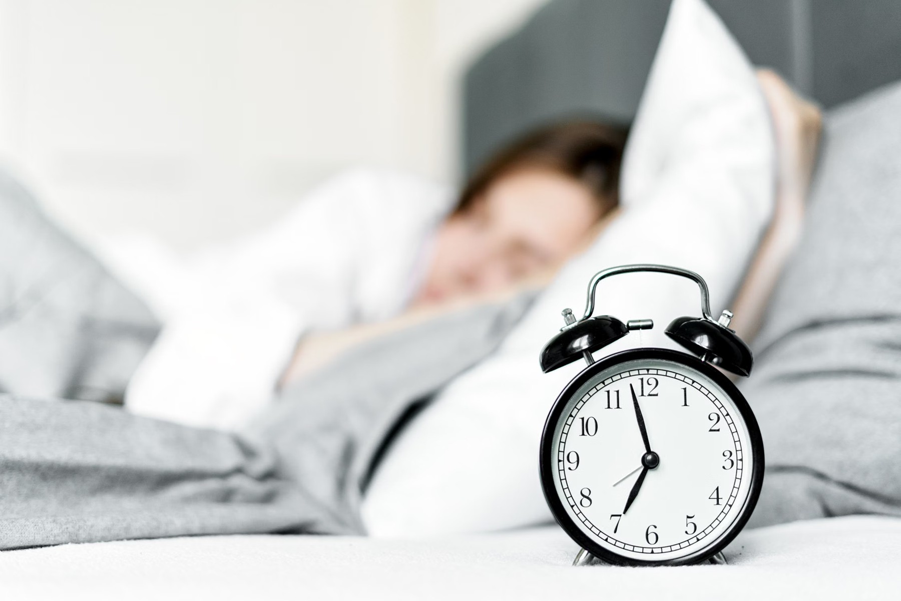 Despertador preto em primeiro plano, com pessoa deitada na cama e desfocada ao fundo.