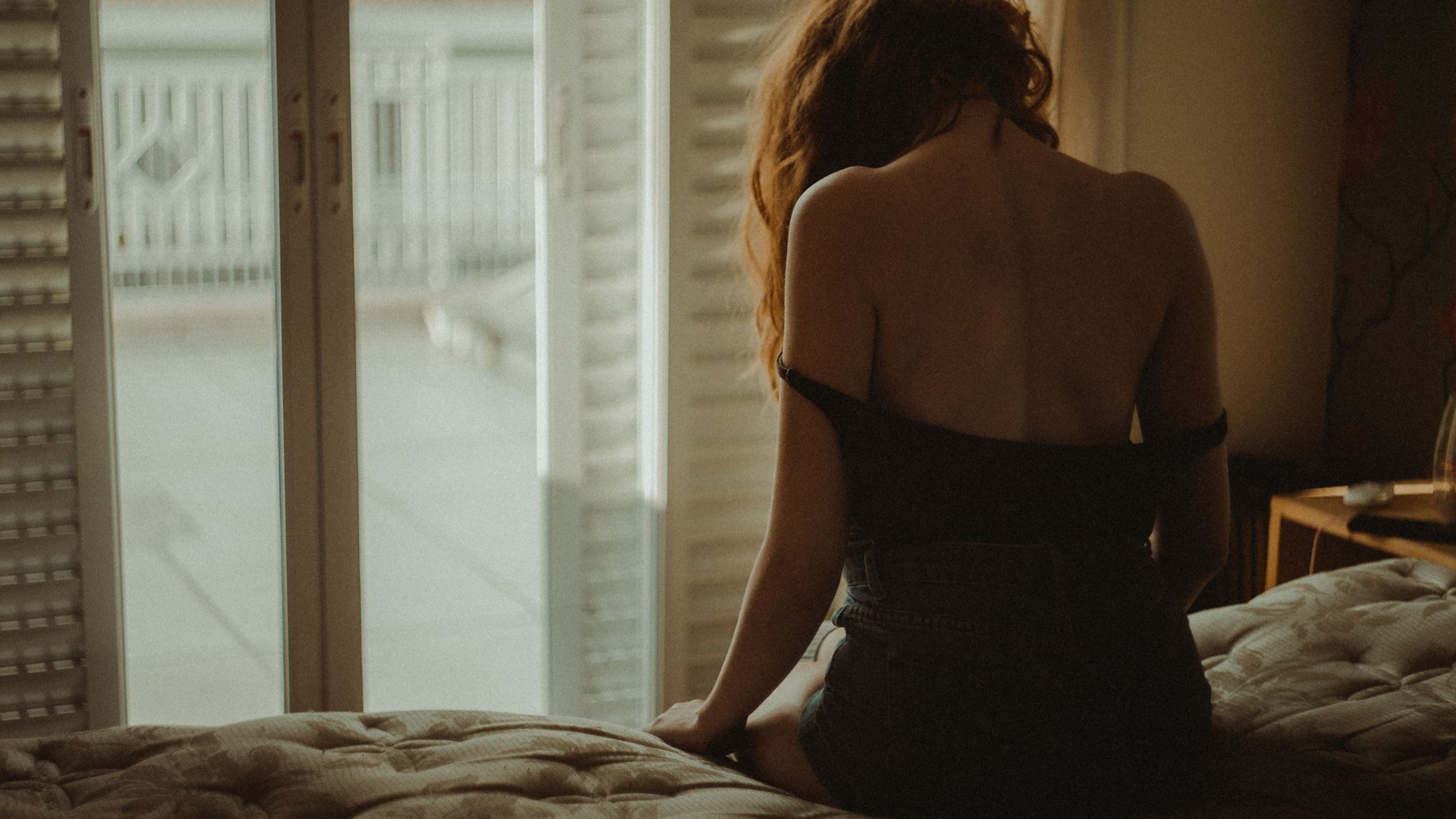 Mulher vista de costas, sentada na cama perto da janela.